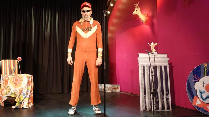 Foto von Daddy R, im ausgewaschenem rostbraunen Onesie, Sonnenbrille, Rapperkette, MCGA Capppy, weiße Socken und Schlappen auf der Bühne des BKA Theaters anlässlich einer Comedy NOW Aufführung