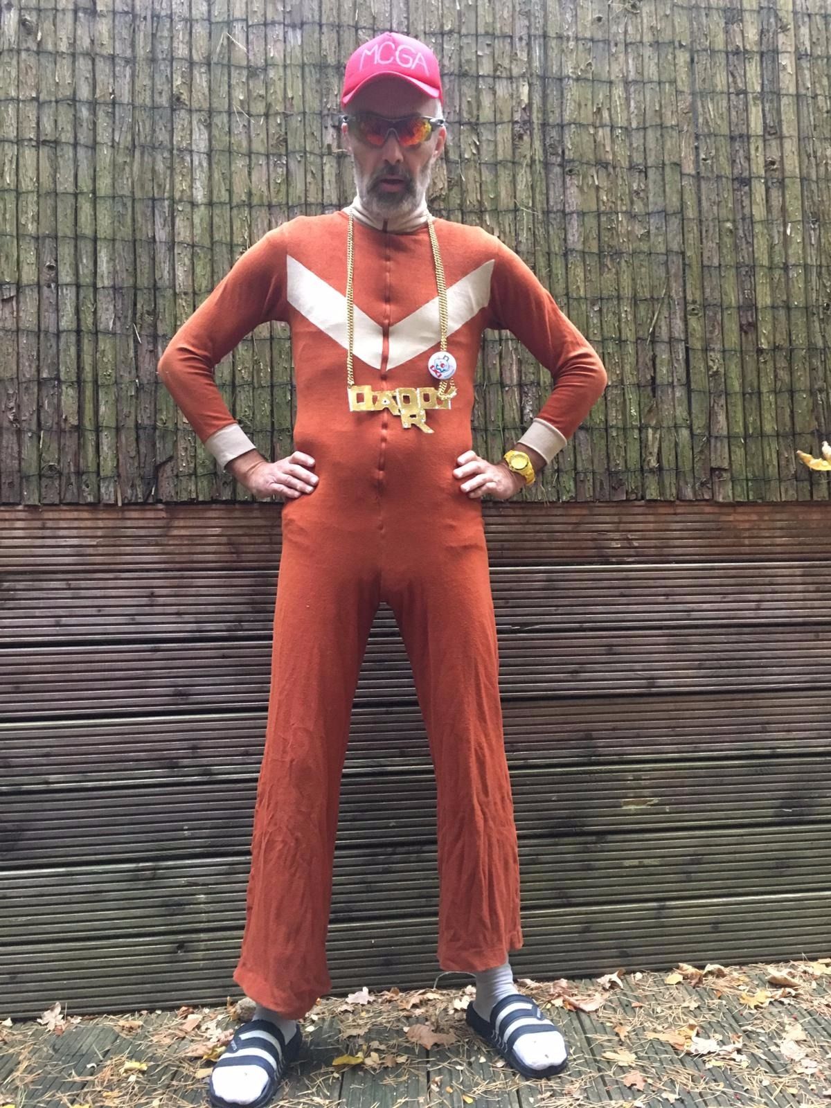 Foto von Daddy R, im ausgewaschenem rostbraunen Onesie, Sonnenbrille, Rapperkette, weiße Socken und Schlappen vor einem Holzzaun