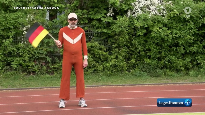 Foto von Daddy R, im ausgewaschenem rostbraunen Onesie, Sonnenbrille, Rapperkette, weiße Socken und Schlappen aus den ARD Tagestehmen mit Deutschlandfahne anlässlich der EM 2016