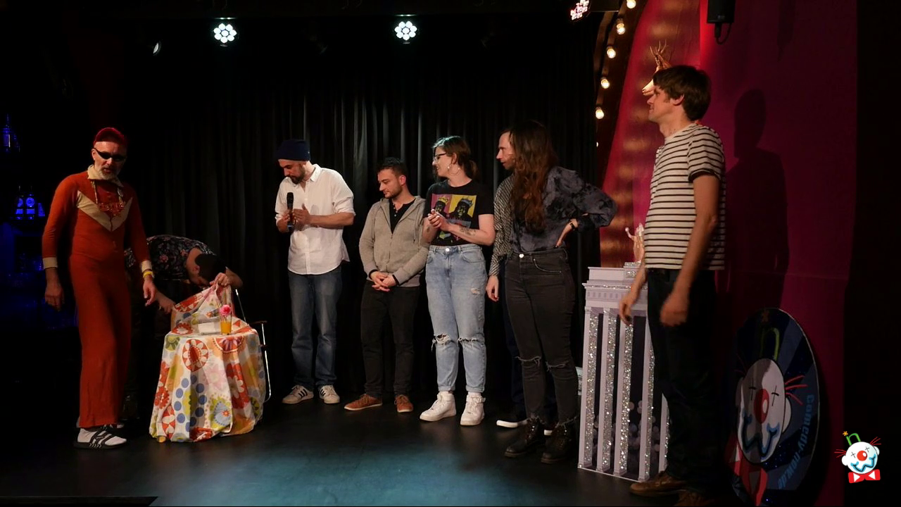 Screenshot von Daddy R, im ausgewaschenem rostbraunen Onesie, Sonnenbrille, Rapperkette, MCGA Capppy, weiße Socken und Schlappen auf der Bühne des BKA Theaters anlässlich einer Comedy NOW Aufführung, inklusive dem Rest des Casts, Thomas Kornmaier, Falk Pyrczek (auf der Suche nach den Proseccos), Franky Fuzz, Lena Beermann, Marc Adams, Vanessa Willi und Niclas Amling, welche am Klatschen sind